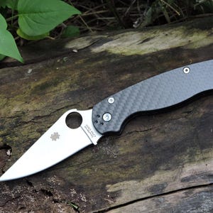 May include: A folding knife with a silver blade and a dark gray carbon fiber handle. The blade has a circular opening and a small logo. The handle has a textured pattern and a pocket clip. The knife is lying on a wooden surface.