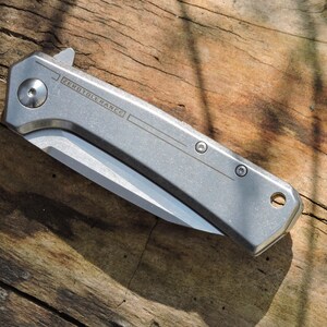 May include: A silver folding knife with a partially exposed blade, resting on a weathered wooden surface. The knife has the text "ZEROTOLERANCE" etched on the side. The blade is silver and the handle is a matte silver color.