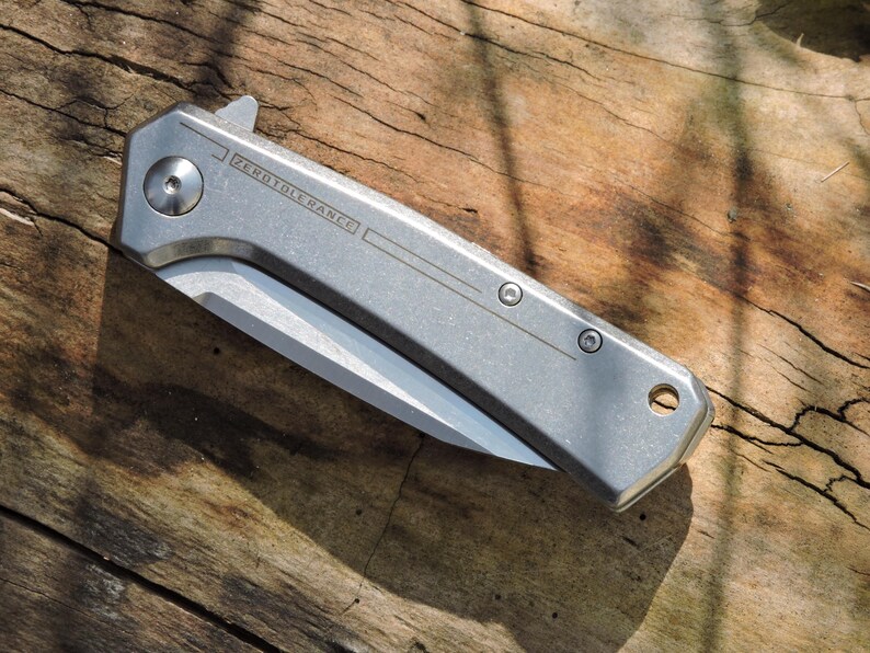 May include: A silver folding knife with a partially exposed blade, resting on a weathered wooden surface. The knife has the text "ZEROTOLERANCE" etched on the side. The blade is silver and the handle is a matte silver color.