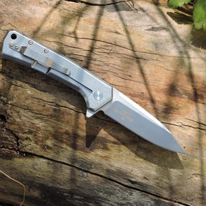 May include: A silver folding knife with a partially serrated blade rests on a weathered wooden surface. The knife features a pocket clip and a small circular button. The blade has text etched on it.