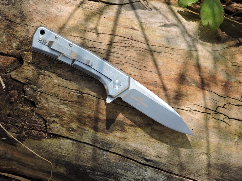 May include: A silver folding knife with a partially serrated blade rests on a weathered wooden surface. The knife features a pocket clip and a small circular button. The blade has text etched on it.