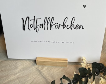 Op de afbeelding: Een witte kaart met de Duitse tekst "Notfallkörbchen" in een handgeschreven lettertype, met een klein hartsymbool. Hieronder staat de tekst "KURZE PAUSE & AB AUF DIE TANZFLÄCHE" gedrukt. De kaart wordt ondersteund door een klein houten blok, met groen en beige blad in de voorgrond.