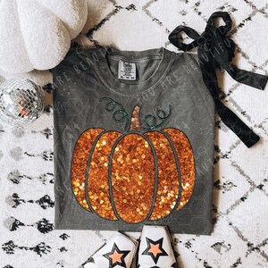 May include: A gray t-shirt with a glittery orange pumpkin design, a black bow, and star-patterned boots. A white plush pumpkin and a disco ball are also present. The shirt has the text "YOU ARE BEAUTIFUL".