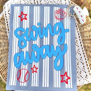 May include: Gray t-shirt with blue text that says "Swing Away". The shirt has white vertical stripes and red baseball-themed graphics, including stars, a baseball, and a glove. A baseball-themed shirt.