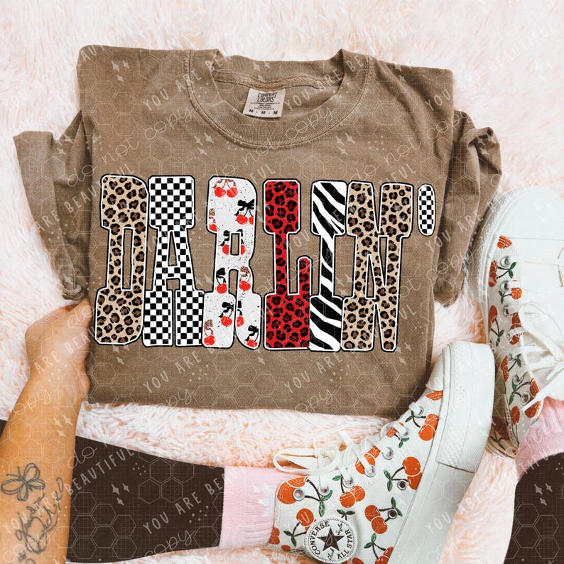 May include: A brown long-sleeve shirt with the word "DARLIN'" in a bold, patterned design. The letters feature leopard print, checkerboard, cherries, and zebra stripes. The shirt is paired with white canvas shoes with cherry graphics.