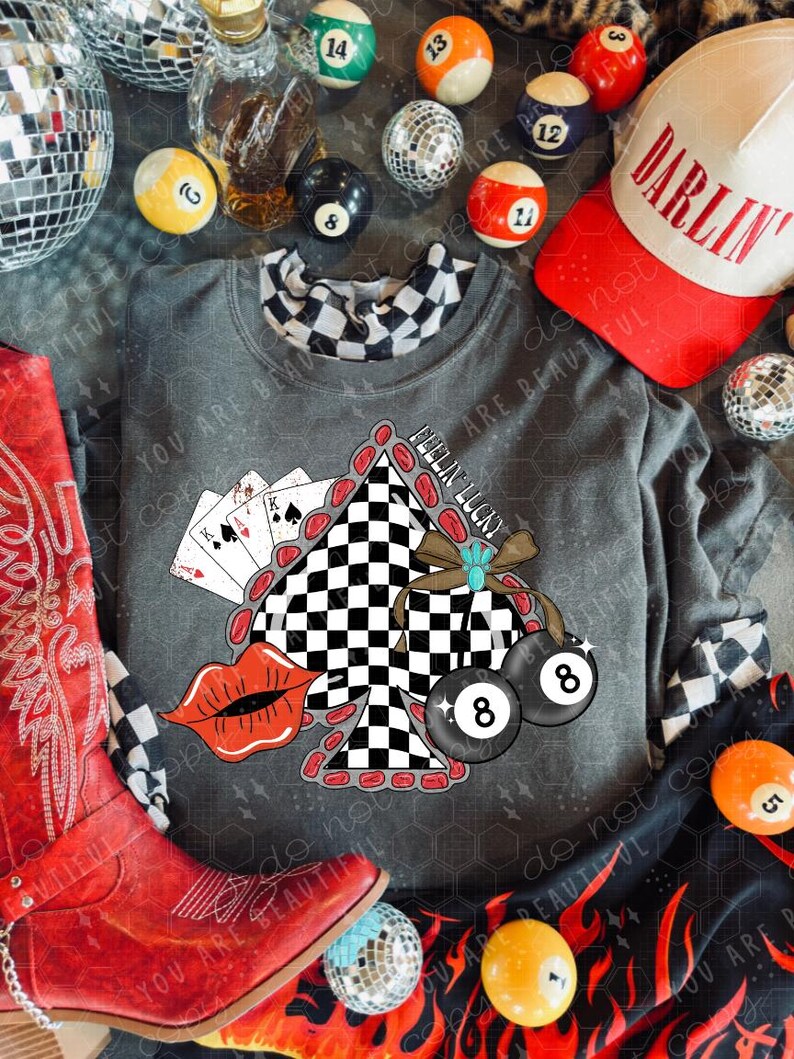 May include: A gray sweatshirt with a graphic featuring playing cards, a checkered design, and two 8-ball pool balls. A red cowboy boot, a baseball cap with "DARLIN" text, and various billiard balls are also visible.