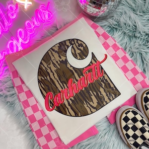 May include: A white t-shirt featuring the Carhartt logo in a brown camouflage pattern with pink lettering. The shirt is folded on a pink and white checkered surface, with a pair of black and white checkered Vans sneakers visible.