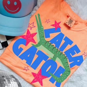 May include: Orange t-shirt with a green alligator graphic and the words "Later Gator" in blue. Pink star accents surround the alligator and text. A light blue trucker hat with a red smiley face is visible.