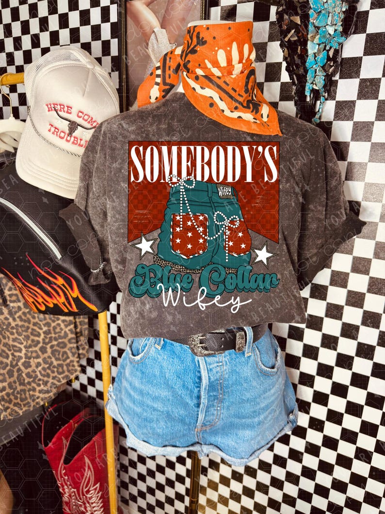 May include: A display featuring a grey t-shirt with a graphic of blue jean shorts and the text "SOMEBODY'S Blue Collar Wifey." The shirt is paired with denim shorts and a black belt. An orange bandana and a white trucker hat are also visible.
