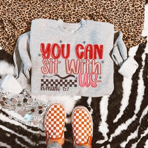 May include: A gray sweatshirt with the text "YOU CAN Sit With Us" in red and pink, with stars and a checkered design. The text "Romans 15:7" is below. The image also shows a pair of orange and white checkered slip-on shoes.