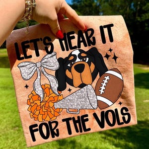 May include: A tan-colored graphic featuring a dog, football, bow, megaphone, and pom-pom. Text reads "LET'S HEAR IT FOR THE VOLS". The dog has black, brown, and white markings. The pom-pom is orange.