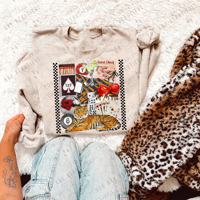 K&ouml;nnte beinhalten: Ein beiger Pullover mit einem grafischen Aufdruck eines Tigers, Spielkarten, W&uuml;rfeln, Kirschen, Streichh&ouml;lzern, einem Cocktail und einer Billardkugel. Die Grafik ist im Collage-Stil mit einem karierten Hintergrund.