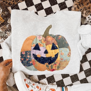 May include: A light gray sweatshirt featuring a patchwork pumpkin design. The pumpkin has a smiling face and is made of various floral and patterned fabrics in shades of orange, green, and purple. The sweatshirt is a casual, comfortable style.