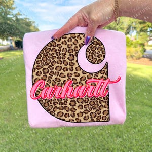 May include: A light pink t-shirt featuring a large, stylized "C" logo filled with a brown leopard print. The word "Carhartt" is written in pink script across the logo. The shirt is held up against a green grassy background.