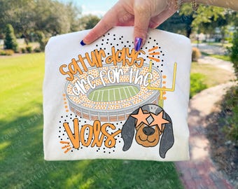 May include: White t-shirt with a cartoon dog wearing star-shaped sunglasses, a football stadium, and the text "Saturdays are for the Vols" in orange. The design includes black polka dots and orange accents.