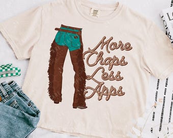 May include: A beige t-shirt with a graphic of a woman wearing blue jeans and brown chaps. The text "More Chaps Less Apps" is written in a rope-like font.