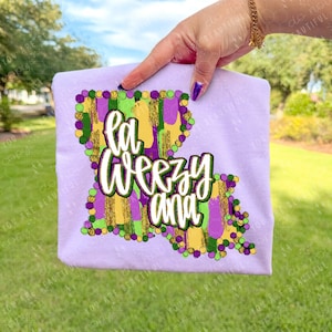 May include: A light purple t-shirt featuring the outline of Louisiana with the words "La Weezy Ana" in a white, green, and purple design. The design includes gold, green, and purple accents, evoking a festive theme.