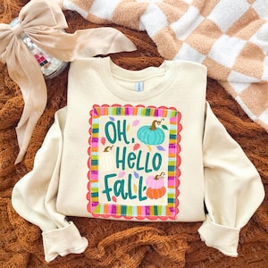 May include: A cream-colored crewneck sweatshirt featuring a central graphic with the text "OH, HELLO FALL" in teal. The design includes illustrations of three pumpkins in teal, white, and orange, surrounded by a colorful, scalloped rectangular border.