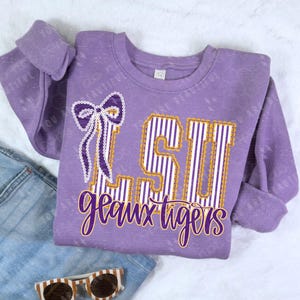 May include: A purple sweatshirt with the letters "LSU" in white and gold stripes, and the words "geaux tigers" in a cursive font. A bow is on the left side of the letters. Sunglasses are at the bottom.