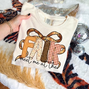 May include: Cream-colored sweatshirt with the word "FAIR" in orange gingham, floral, and leopard print letters, and a brown bow. The text "meet me at the" is below the word "FAIR". The sweatshirt is on a patterned surface.