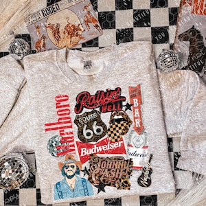 May include: A gray sweatshirt with a graphic design featuring a variety of vintage-inspired imagery, including a Route 66 sign, a Budweiser logo, a guitar, a pair of lips, and the text "Raisin' Hell" and "Marlboro".