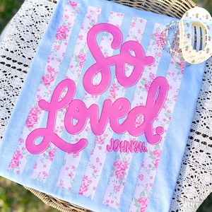 May include: A light blue blanket with pink floral stripes and the words "So Loved" in large pink script. The text "John 3:16" is below the word "Loved". A floral hair clip and bracelets are on the blanket.