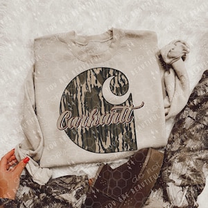 May include: A beige sweatshirt featuring the Carhartt logo in a camouflage pattern. The brand name is written in a cursive font. The sweatshirt is paired with camouflage pants and brown boots.