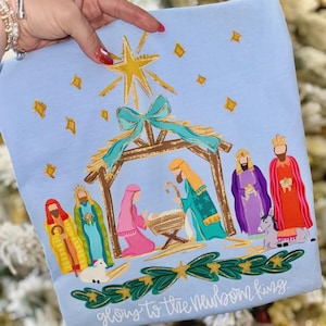 May include: A light blue Christmas-themed graphic featuring a nativity scene with a star, stable, and figures in vibrant colors. The text "glory to the newborn king" is at the bottom. The design is on a soft fabric.