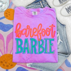 May include: A lilac-colored t-shirt with the words "barefoot BARBIE" in orange and green script. The shirt is laid on top of a pair of blue jeans and white sneakers. A disco ball and floral pattern are in the background.