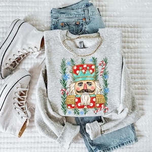May include: A light gray crewneck sweatshirt featuring a graphic of a nutcracker with a teal crown, red coat, and gold epaulets, surrounded by green pine branches and candy canes, paired with blue denim jeans and white high-top sneakers.