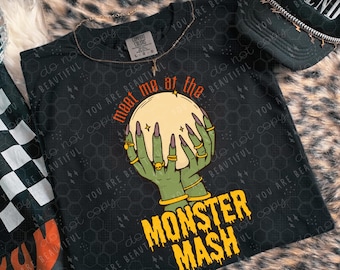 Może przedstawiać: Czarny t-shirt z grafiką zielonych dłoni trzymających kryształową kulę, z napisem "meet me at the" i "MONSTER MASH". Widoczna jest również czarna czapka z napisem "LEGEND".