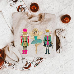 May include: A beige sweatshirt featuring colorful illustrations of a nutcracker, a fairy, and a mouse king. The sweatshirt is decorated with Christmas ornaments and a festive patterned fabric.