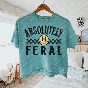 May include: A teal blue cropped t-shirt with the text "ABSOLUTELY FERAL" in black. A yellow smiley face with black eyes is above the text. A black and white checkered pattern is below the text.