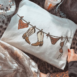 May include: A white sweatshirt featuring a graphic print of a clothesline with a red bandana, two brown and white cowboy boots, and a brown striped bikini set hanging from it. The design is centered on the front of the garment.