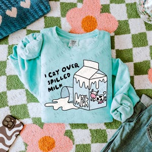 May include: A light blue sweatshirt with a white graphic of a spilled carton of milk and the text "I cry over spilled milk".