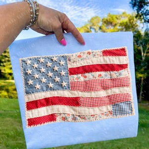 May include: A handmade American flag made of fabric patches in red, white, and blue. The flag features stars and stripes, with a light blue background. The flag is held up against a backdrop of a blue sky and green grass.