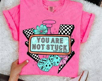 May include: Pink t-shirt with a retro graphic design featuring a checkered pattern, dice, flowers, and a sign that reads "You are not stuck. Don't look back."