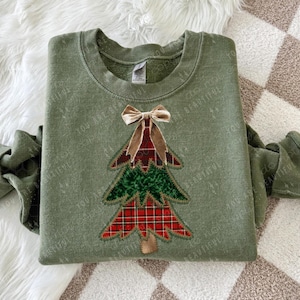 May include: An olive green sweatshirt featuring a Christmas tree design. The tree is made of red plaid and green fabric, with a brown trunk and a tan velvet bow at the top. The words "YOU ARE BEAUTIFUL" are faintly printed on the sweatshirt.