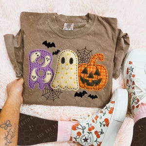 May include: Brown t-shirt with a Halloween-themed design. The design includes the word "BOO" in purple, a ghost, and a jack-o'-lantern. Black bats and spiderwebs complete the festive look. The shirt is paired with white sneakers.