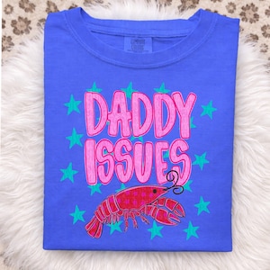 May include: A blue t-shirt with the words "DADDY ISSUES" in pink, surrounded by teal stars. A red lobster graphic is below the text. The shirt is made of Comfort Colors material.