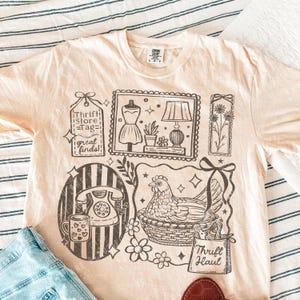 May include: A light peach-colored t-shirt with a graphic design featuring a telephone, dress form, lamp, flowers, and a chicken in a basket. The text includes "Thrift Store = Tag = great finds!" and "Thrift Haul."