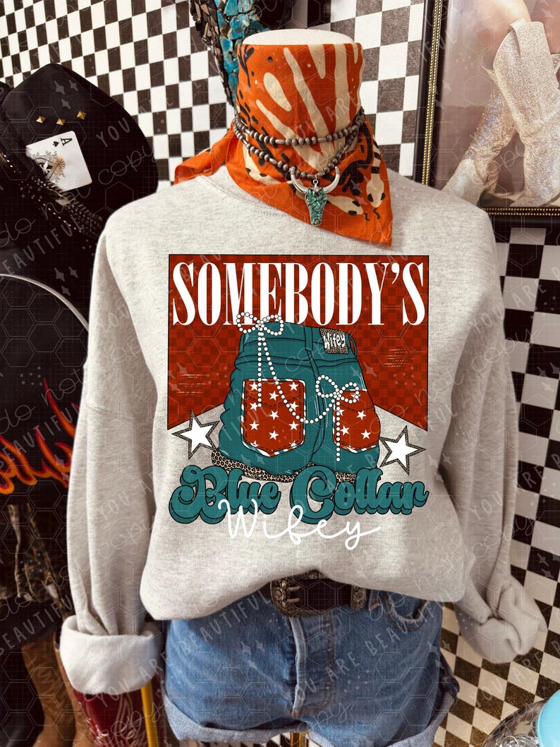 May include: A light grey sweatshirt with a graphic design of blue jeans with red and white stars, the words "SOMEBODY'S" and "Blue Collar Wifey". The sweatshirt is worn with denim shorts and a brown belt.