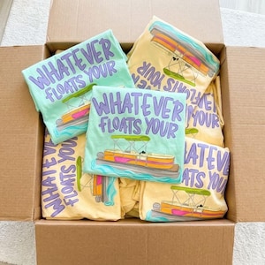 May include: A cardboard box filled with several t-shirts. The shirts are light green and yellow, each featuring a graphic of a pontoon boat and the text "WHATEVER FLOATS YOUR". The shirts are folded and stacked.