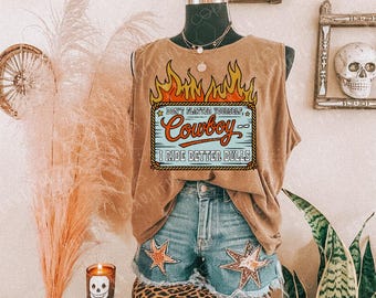 May include: A tan tank top with a graphic that reads "Don't flatter yourself, Cowboy, I ride better bulls" in a flame design. The tank top is paired with denim shorts featuring star patches. The outfit is displayed on a mannequin.