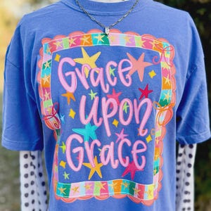 May include: A blue t-shirt with the words "Grace Upon Grace" in pink script, surrounded by colorful stars and a decorative border. The shirt has short sleeves and a crew neck. A silver necklace is visible.