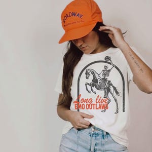 May include: White t-shirt featuring a skeleton riding a skeletal horse, with the text "Long Live Emo Outlaws" in red. The person in the image is wearing an orange baseball cap with the word "Roadway" on it.