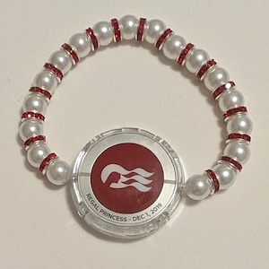 May include: A pearl bracelet with red rhinestone accents and a circular charm. The charm has a red background with a white logo and the text "REGAL PRINCESS - DEC 1, 2019". The bracelet is made of white faux pearls.