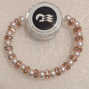 May include: A circular bracelet composed of alternating white faux pearls and rose gold-toned spacers embedded with clear crystals. Positioned above the bracelet is a round, clear plastic container with a black lid featuring a white logo and the text SKY PRINCESS - AUG 8, 2026.