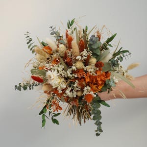 May include: A vibrant dried flower bouquet with a mix of textures and hues. The arrangement showcases orange, white, and green dried flowers and grasses. The bouquet is held in a hand, set against a plain backdrop, creating a natural aesthetic.