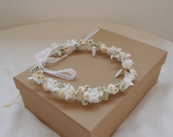 May include: A delicate floral crown featuring white and cream-colored flowers and greenery. The crown is adorned with a white lace ribbon. The floral arrangement is set on a brown cardboard box.